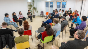 eine Diskussionrunde in einem Raum, vorne sitzen vier Personen auf dem Podium, das Publikum sitzt auf bunten Stühlen